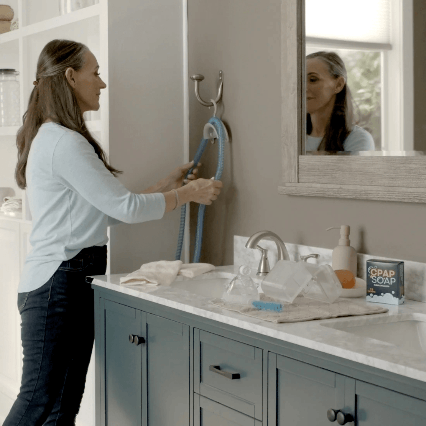 Woman draping a CPAP hose over the CPAP Soap Hose Hanger mounted on a towel rack, with CPAP mask and accessories laid out on the counter