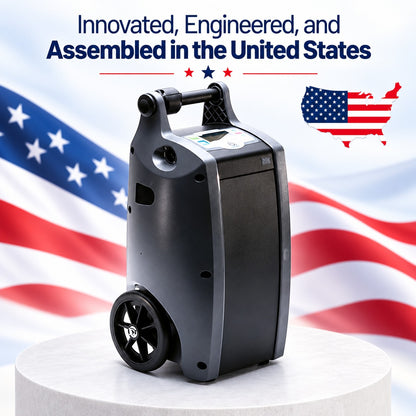 A dark gray O2 Concepts Oxilfe Independence with a handle and front control panel, displayed on a white pedestal against a blurred American flag background. Text at the top reads 'Innovated, Engineered, and Assembled in the United States' with a small U.