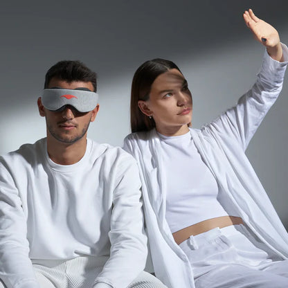 A man and woman in bed together. The man is wearing the manta sleep mask and the woman is putting up her hand to block out the light. 